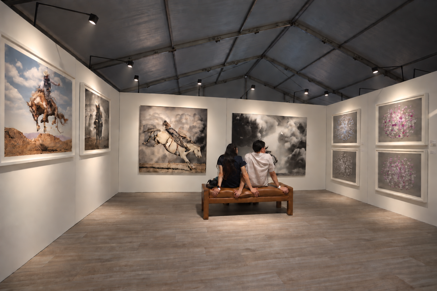 Two visitors seated on a brown bench in a tented gallery, surrounded by illuminated white walls displaying cowboy photographs and floral artwork.